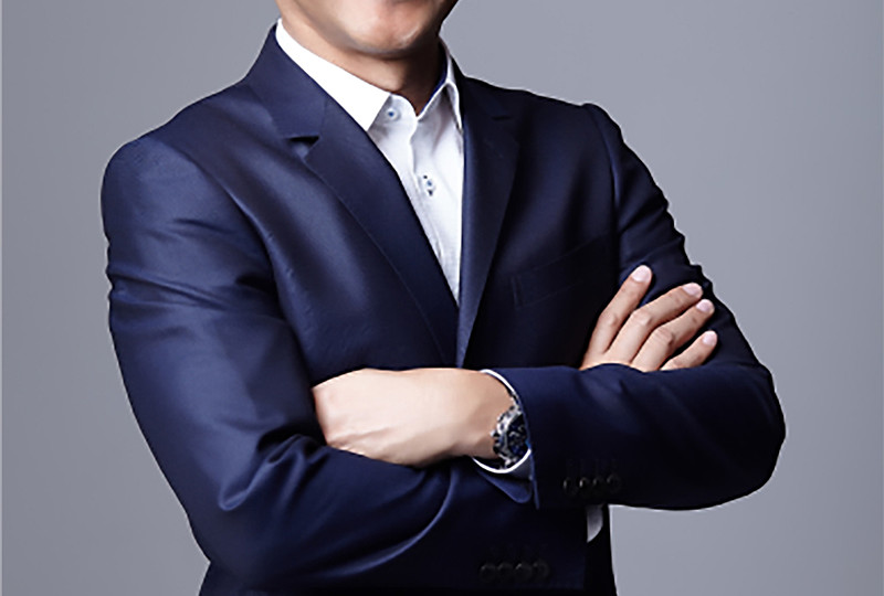 A smiling middle-aged man with dark hair and glasses stands in front of a neutral gray background. He is wearing a dark blue suit with a white shirt and has his arms crossed. The person in the image is Touchawatt Techamongkoljit, CEO of the Thai plastics manufacturer Royce Universal Co. Ltd.