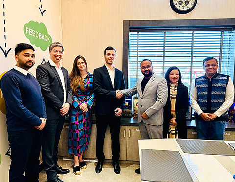 A group of seven people is posing for a photo in a modern office setting. In the center, Leander Huemer from Starlinger (left) and Nitish Goel, CMD of Lakhdatar International Pvt. Ltd. (right), are smiling and shaking hands. Both are wearing suits—Huemer in black, Goel in grey. Behind them, large windows with blinds and several product packages on a sideboard are visible. On the left wall, graphic elements include the word “FEEDBACK.” Everyone appears friendly and professional.