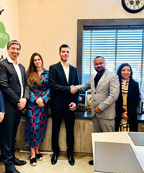 Eine Gruppe von sieben Personen posiert für ein Foto in einem modernen Büro. Zwei Männer in der Mitte, Leander Huemer von Starlinger (links) und Nitish Goel, CMD von Lakhdatar International Pvt. Ltd. (rechts), geben sich lächelnd die Hand. Beide tragen Anzüge, Huemer in Schwarz, Goel in Grau. Im Hintergrund sind große Fenster mit Jalousien sowie Produktverpackungen auf einem Sideboard zu sehen. An der Wand links sind grafische Elemente mit dem Wort "FEEDBACK" sichtbar. Alle Personen wirken freundlich und professionell.