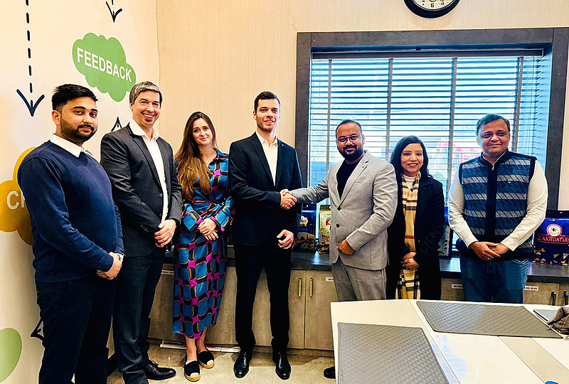 Eine Gruppe von sieben Personen posiert für ein Foto in einem modernen Büro. Zwei Männer in der Mitte, Leander Huemer von Starlinger (links) und Nitish Goel, CMD von Lakhdatar International Pvt. Ltd. (rechts), geben sich lächelnd die Hand. Beide tragen Anzüge, Huemer in Schwarz, Goel in Grau. Im Hintergrund sind große Fenster mit Jalousien sowie Produktverpackungen auf einem Sideboard zu sehen. An der Wand links sind grafische Elemente mit dem Wort "FEEDBACK" sichtbar. Alle Personen wirken freundlich und professionell.