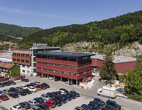 Luftaufnahme des Fabriksgeländes der Firma Starlinger in einer grünen, hügeligen Landschaft. Das Hauptgebäude ist ein modernes, mehrstöckiges Bauwerk mit roter Fassade und großen Fenstern. Auf dem Firmenparkplatz stehen zahlreiche Autos. Im Hintergrund sind bewaldete Hügel und Felsen zu sehen, die eine malerische Kulisse für den Standort bieten.