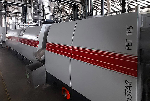Eine großdimensionierte PET-Recyclinganlage vom Typ recoSTAR PET 165 der Firma Starlinger steht in einer Industriehalle. Die Maschine ist mit roten Streifen versehen und von Rohrleitungen, Plattformen und Stahlkonstruktionen umgeben. Es handelt sich um eine Bottle-to-Bottle-Recyclinganlage, installiert im Werk des thailändischen Kunststoffherstellers Royce Universal Co. Ltd.