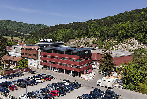 Luftaufnahme des Fabriksgeländes der Firma Starlinger in einer grünen, hügeligen Landschaft. Das Hauptgebäude ist ein modernes, mehrstöckiges Bauwerk mit roter Fassade und großen Fenstern. Auf dem Firmenparkplatz stehen zahlreiche Autos. Im Hintergrund sind bewaldete Hügel und Felsen zu sehen, die eine malerische Kulisse für den Standort bieten.